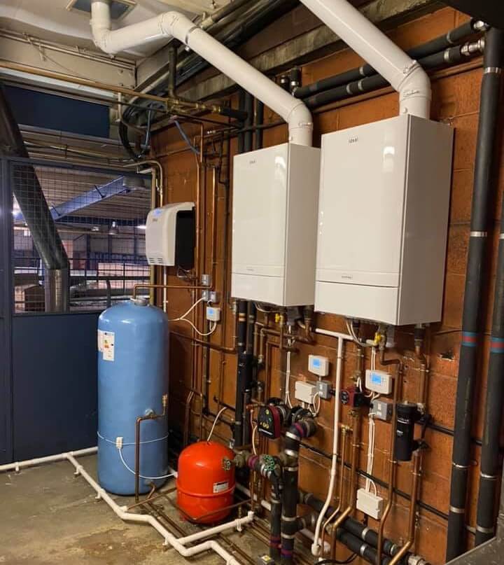 Commercial heating system installation featuring two white boilers, blue expansion tank, and various pipes and valves on a wall, illustrating JA Plumbing and Heating's expertise in heating solutions for businesses.